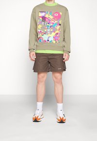 Olivgrön sweatshirt med livfullt grafiskt tryck, lagd över en ljusgrön skjorta. Bruna cargoshorts med sidofickor, sportskor med orange detaljer.