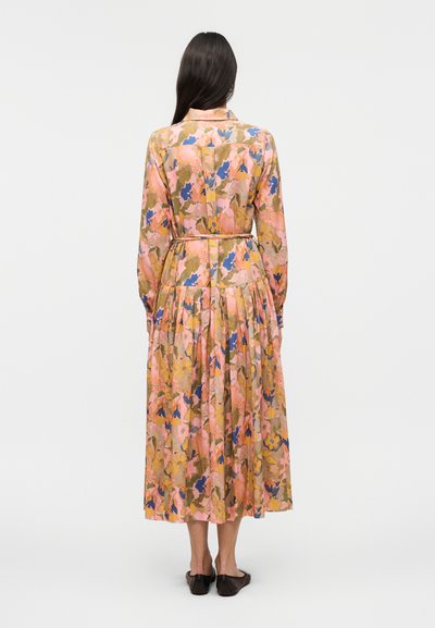 Hosszú sötét hajú nő, aki egy öves, hosszú ujjú, bokáig érő virágmintás ruhát visel, mely rózsaszín, kék és sárga mintákkal díszített, fehér háttér előtt áll.