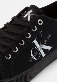 Zapatillas de lona negras con diseño de cordones, punta redonda, ojetes plateados y suela de goma. Presenta el logotipo blanco impreso "CK" y "Calvin Klein Jeans".