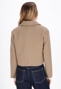 Giacca beige in misto lana, tagliata, con colletto a revers, caratterizzata da una texture liscia e cuciture sartoriali, abbinata a jeans denim scuri.