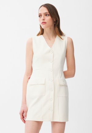 Kobieta o brązowych włosach do ramion, ubrana w białą, bezrękawową sukienkę zapinaną na guziki z przednimi klapkowymi kieszeniami, patrząca na bok.