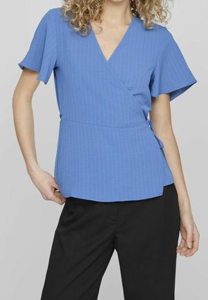 Vrouw die een blauwe korte mouwen overslagblouse met subtiele verticale strepen en zwarte broek draagt, staand tegen een effen achtergrond.