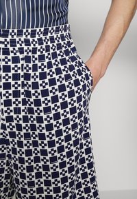 Main d'homme dans la poche d'un short à carreaux bleu marine et blanc avec taille élastique et motif texte "Sandro".