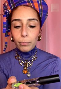 Donna con dolcevita viola e fazzoletto a quadri in testa tiene una bottiglia di profumo trasparente con tappo nero su sfondo rosa.