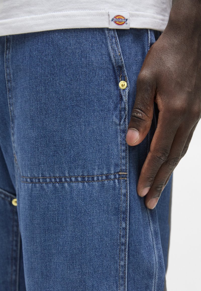 Spijkerbroek in medium blauw met een soepele textuur, voorzien van gouden hardware knopen en zichtbare stiksels. Dickies logo label zichtbaar.