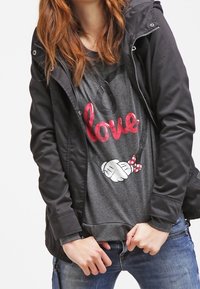 Giacca nera con zip sopra una maglietta grigia che presenta una grafica "love" in rosso e mani in cartoon con un fiocco a pois. Indossata con jeans blu.