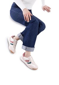 Las zapatillas presentan un upper beige con franjas rojas y verdes, acentos blancos y una suela de goma. Combinadas con unos jeans azul oscuro con puños doblados.