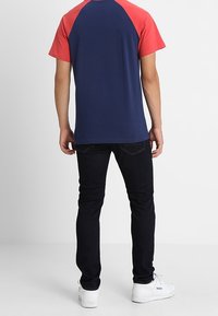 Krátký rukáv tričko s námořnickým tělem a červenými raglánovými rukávy, v kombinaci s tmavými džíny a bílými teniskami, pohled zezadu.