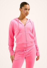 Rožinė velūro vos priekyje užtraukiama striukė turi priekinius kišenes, auksinį užtrauktuką ir raiščius. Minkšta medžiaga puikiai dera su rožiniais džinsais.