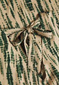 Tela beige con patrón abstracto en verde; incluye una característica de lazo central atado. Material texturizado y suave con un diseño fluido.