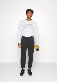 Maglietta grigia chiara a maniche lunghe con logo circolare nero, abbinata a pantaloni della tuta neri, sneaker nere e in mano una bottiglia d'acqua gialla.