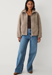 Veste en polaire beige avec col montant, fermeture éclair et deux poches latérales. Associée à un jean bleu à jambes larges et des bottines marron.
