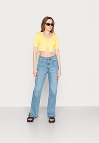 Top cropped amarelo canelado com mangas curtas e um detalhe recortado à frente, combinado com jeans de perna reta azul claro de cintura alta e sandálias pretas.