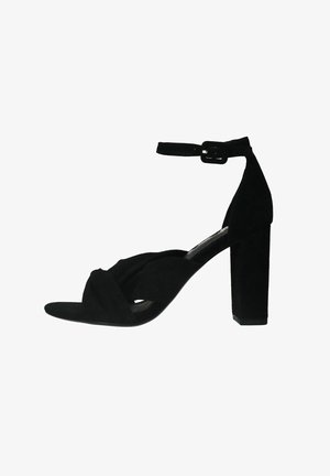 Zwarte suède hoge hak sandaal met een gedraaide ontwerp op de teen, enkelband met gesp en een dikke blokhak voor stabiliteit.