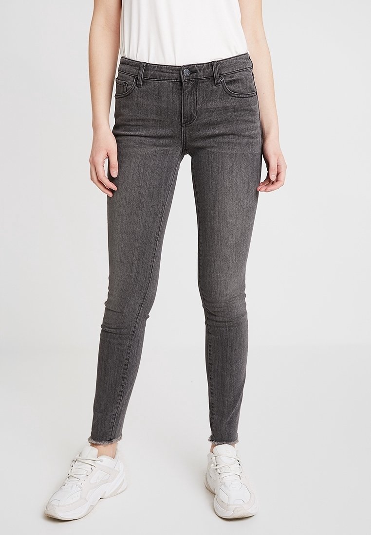 Personne portant un jean skinny gris foncé avec des ourlets effilochés, des baskets blanches et un t-shirt blanc rentré, debout sur un fond uni.