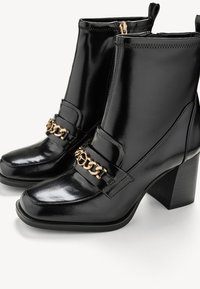 Botas de tobillo de charol negro con puntera cuadrada, acento de cadena dorada y tacón grueso. Presentan un cierre de cremallera en la parte trasera.