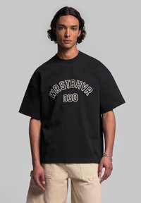 Černé oversized tričko s bílým vyšitým textem "WRSTBHV" a "030" na přední straně. Měkká bavlněná tkanina a krátké rukávy.