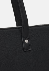 Coin du sac fourre-tout en cuir noir avec une surface texturée, doté d'un bouton-pression argenté et d'une bandoulière noire robuste. Détail de fermeture éclair visible.