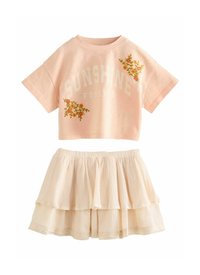 Rosa Baumwoll-Crop-T-Shirt mit dem Aufdruck "SUNSHINE FOREVER" und floraler Stickerei, kombiniert mit einem geschichteten cremefarbenen Rock mit strukturiertem Design und Spitzenbesatz.