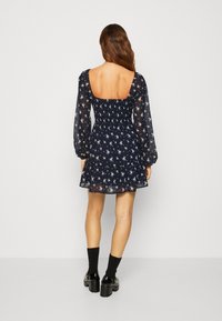 Marinblommig klänning med långa genomskinliga ärmar, fyrkantig rygg, smockad midja och volangnederdel. Bärs med svarta strumpor och kraftiga skor.