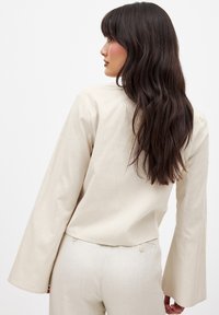 Beige langærmet top med flagrende ærmer, der har en enkel rund halsudskæring. Stoffet ser struktureret ud, med en afslappet pasform bagpå.