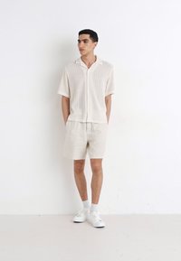 Crème gebreid shirt met korte mouwen en knopen aan de voorkant, gecombineerd met lichtbeige shorts. Draagt witte sneakers en sokken, tegen een witte achtergrond.