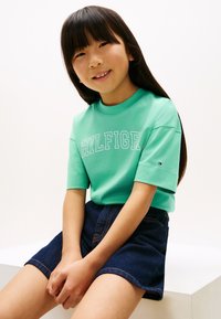 Ανοιχτό πράσινο βαμβακερό t-shirt με κοντά μανίκια και το κείμενο "HILFIGER"· συνδυασμένο με σκούρα τζιν φούστα με πορτοκαλί ραφές.