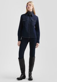 Camicia di jeans blu scuro con due tasche sul petto, chiusura con bottoni e maniche lunghe. Indossata con jeans skinny blu scuro e stivali neri in pelle al ginocchio.