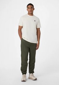 Kremowa koszulka z krótkim rękawem z małym logo, zestawiona z zielonymi joggerami. Joggery mają mankiety przy kostkach i są noszone z jasnymi tenisówkami.