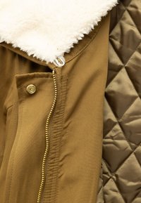 Bruine buitenkleding met een crème nepbontkraag, voorzien van een gouden ritssluiting en drukknoopsluiting, met een doorgestikte binnenvoering.