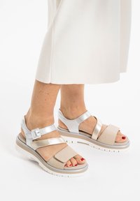 Lettrede sandaler lavet af beige ruskind og sølv syntetisk materiale. Har krydset remme og en tykkere hvid gummisål.