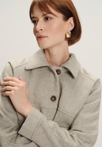 Cappotto grigio chiaro con motivo a scacchi, dotato di colletto e bottoni marroni, realizzato in un tessuto morbido e dalla vestibilità sartoriale.