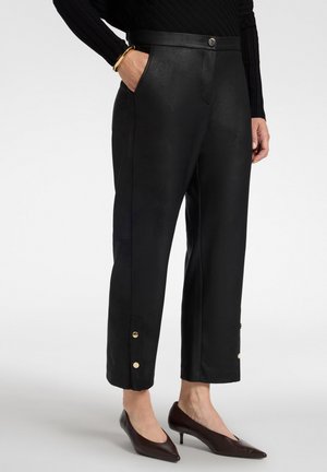 Frau trägt schwarze knöchellange Hose mit goldenen Knöpfen an den Manschetten, schwarzen gerippten Oberteil, goldenen Armreif und dunkelbraune Absatzschuhe.
