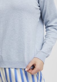Maglione a maglia azzurro chiaro con polsini e orlo a coste; presenta una texture liscia e una vestibilità comoda, abbinato a pantaloni a righe blu e bianchi.
