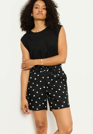 Femme aux cheveux bouclés portant un haut noir sans manches et un short noir à pois blancs, debout les bras croisés devant un fond uni.