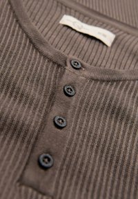 Shirt henley a coste marrone con cinque bottoni neri, scollatura rotonda e un'etichetta con la lettera "M". La texture è morbida e uniforme.