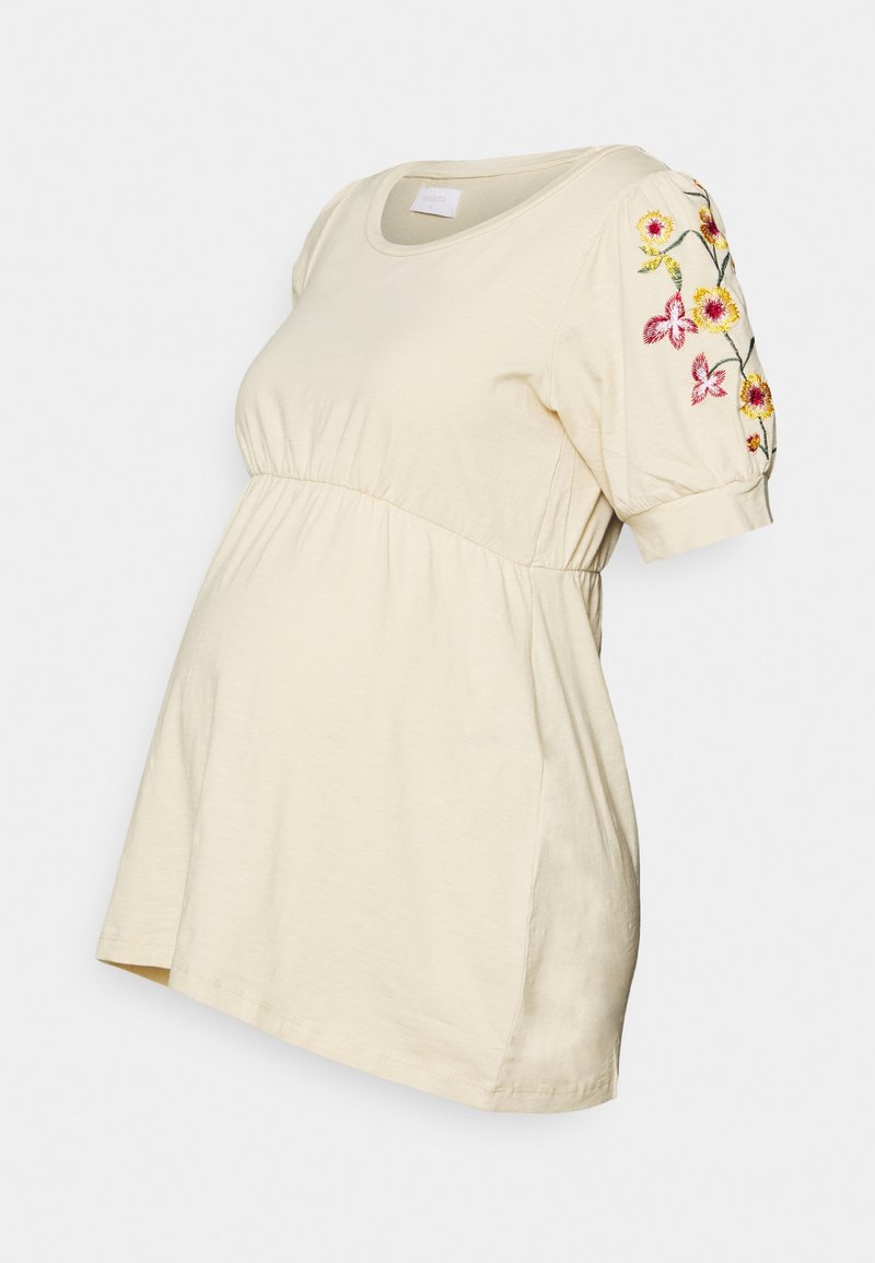 Lyse beige graviditetst-shirt med korte pufferærmer, indsnøret talje og farverig blomstret broderi på ærmerne, lavet af blødt bomuldsmateriale.