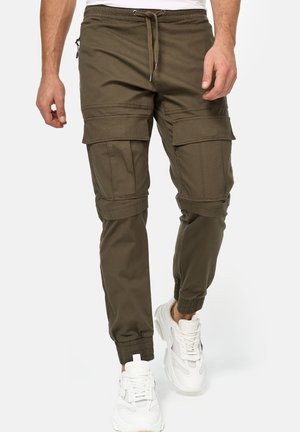 Mężczyzna noszący oliwkowe spodnie bojówki typu jogger z ściągaczem w pasie i elastycznymi mankietami przy kostkach, w połączeniu z białymi trampkami.