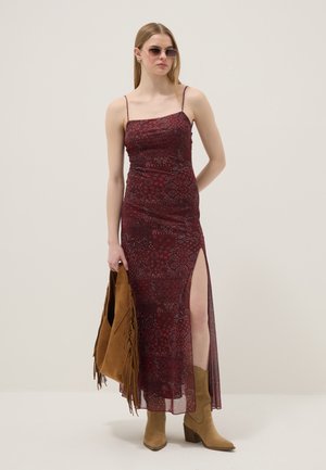 Donna che indossa un vestito slip floreale color bordeaux con spacco sulla coscia, stivali da cowboy color cuoio, occhiali da sole e tiene una borsa a frange color cuoio.