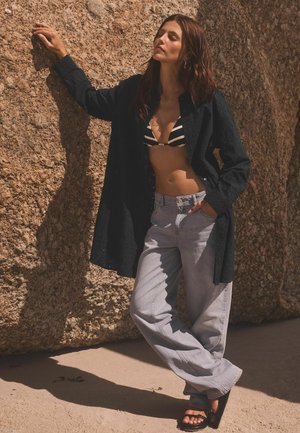 Schwarzes Langarmshirt, gestreifter Bikinioberteil, hellgraue weite Jeans und schwarze Sandalen, posiert vor einem felsigen Hintergrund.