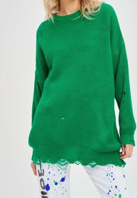 Femme portant un pull vert oversize en maille avec des bords usés et un pantalon blanc éclaboussé de peinture bleue et verte.