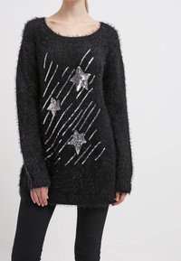 Jersey negro y peludo con un escote amplio y mangas largas. Presenta motivos en forma de estrella de lentejuelas plateadas y detalles de rayas diagonales.