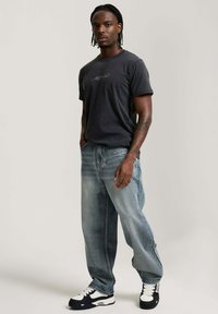 Man with braids wearing a dark graphic t-shirt, loose faded jeans, and black-and-white sneakers posing with hands in pockets.