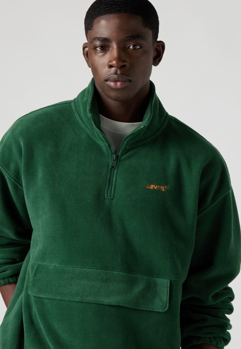 Grön fleecetröja med halvdragkedja, utrustad med en framficka och broderad orange logotyp. Mjuk textur och avslappnad passform.