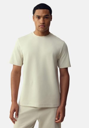 Jeune homme debout, face à l’avant, portant un t-shirt uni beige clair à manches courtes et un pantalon assorti sur fond blanc.