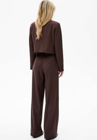 Brązowa krótka marynarka z długimi rękawami, zestawiona z spodniami o wysokim stanie i szerokich nogawkach. Materiał wydaje się gładki, z minimalnymi detalami.