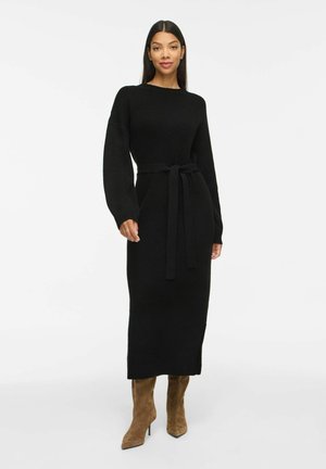 Femme portant une longue robe noire en maille avec une ceinture à la taille et des bottes marron en daim à talons, debout sur un fond blanc uni.