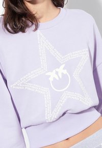 Lila collegepaita, jossa on valkoinen tähtikuosi ja toistuva tekstilogo. Kuvio sisältää pyöreän yksityiskohdan, jossa on kaksi tyyliteltyä lintua.