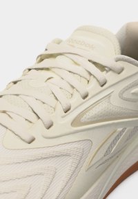 Κοντινό πλάνο εκρού αθλητικού παπουτσιού Reebok που δείχνει υφασμάτινη υφή, κορδόνια και λογότυπο στη γλώσσα και την πλευρά.