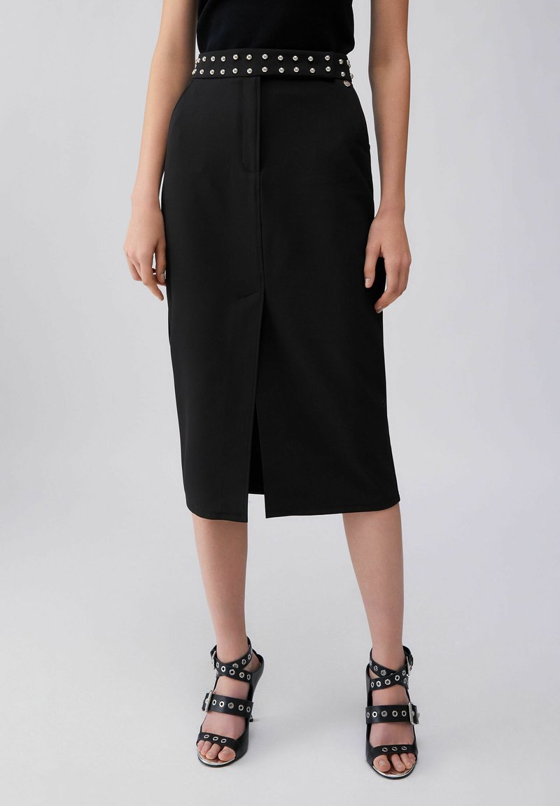 Naine, kes kannab musta midi-seelikut esilõhikuga ja naelutatud vööd, kombineeritud mustade kontsakingadega, millel on mitu rihma.